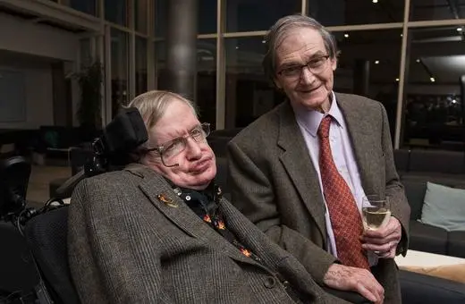 Stephen Hawking sits in a wheelchair, smiling, next to a man holding a glass of champagne, both in a modern indoor setting.