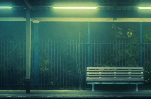A dimly lit, empty bench at a train station, surrounded by mist and foliage, conveying a quiet, eerie atmosphere.
