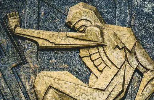 A golden relief sculpture of a worker in a dynamic pose, showcasing muscular arms and industrial elements, representing strength and perseverance.