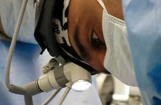 A focused surgeon wearing a surgical mask and headlamp, leaning over a patient during an operation, surrounded by surgical drapes and equipment.