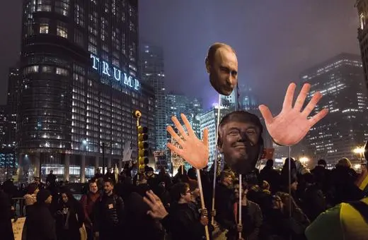 Protesters hold large cutouts of Donald Trump and Vladimir Putin while demonstrating at night in Chicago, with the Trump Tower illuminated in the background.