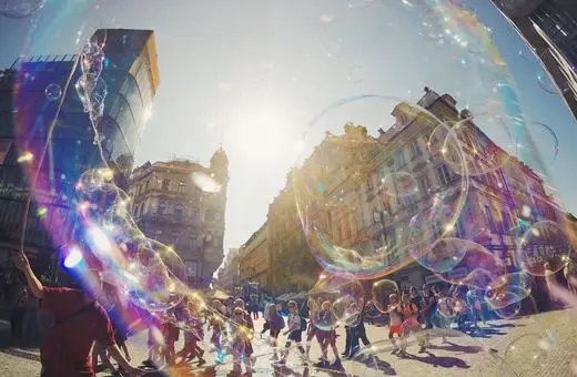 A vibrant street scene with people walking under a clear sky, surrounded by large bubbles reflecting sunlight and historic buildings in the background.