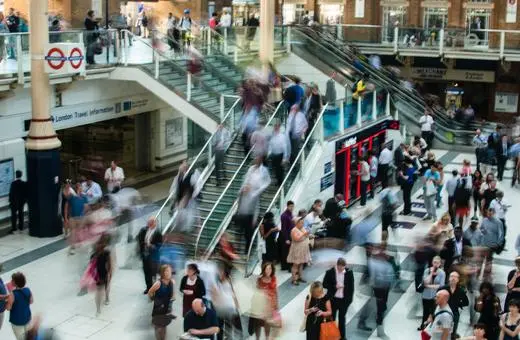 A busy railway station filled with commuters in motion, featuring escalators, signage, and diverse groups of people engaged in various activities.