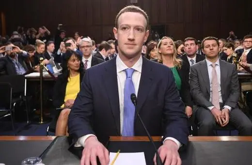 Mark Zuckerberg sitting before a congressional hearing, with a crowd of onlookers and journalists in the background.