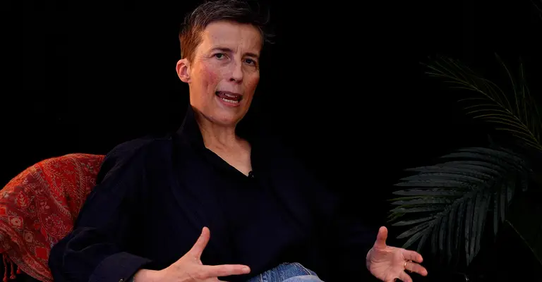 A person with short hair is seated, gesturing as they speak, against a dark backdrop with a plant and a patterned fabric behind them.
